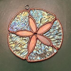 May include: A stained glass sand dollar suncatcher with a copper-colored frame. The center features a flower design with five petals. The glass has a textured, iridescent finish with shades of blue, green, and yellow. A small metal loop is attached for hanging.