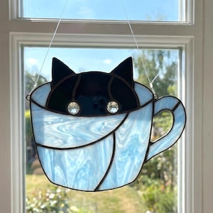 May include: Stained glass cat in a teacup. The cat's head is black, with two crystal eyes. The teacup is light blue with a handle. The glass is leaded with black lines. The piece is hanging in front of a window.
