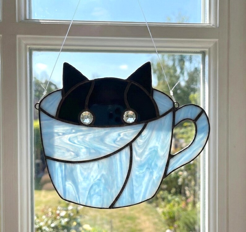 May include: Stained glass cat in a teacup. The cat's head is black, with two crystal eyes. The teacup is light blue with a handle. The glass is leaded with black lines. The piece is hanging in front of a window.