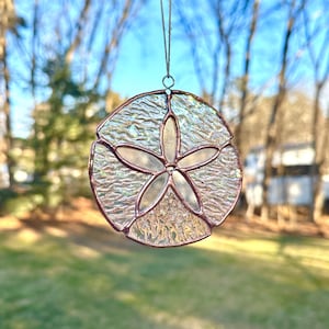 May include: A stained glass sand dollar suncatcher with a textured, iridescent finish. The sand dollar design features a five-pointed star in the centre, surrounded by a circular frame. The suncatcher is suspended by a thin string.
