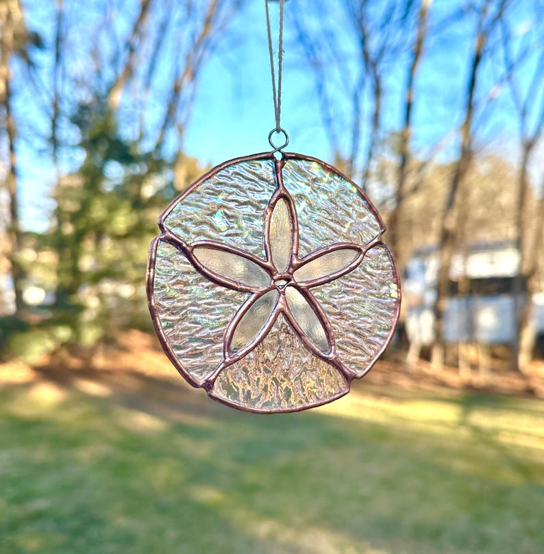 May include: A stained glass sand dollar suncatcher with a textured, iridescent finish. The sand dollar design features a five-pointed star in the centre, surrounded by a circular frame. The suncatcher is suspended by a thin string.