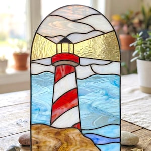 Puede incluir: Panel de vidrieras que representa un faro con rayas rojas y blancas, una parte superior roja y una luz dorada. El faro se encuentra sobre una masa de tierra marrón y beige, con agua y cielo azules y blancos. El panel tiene una parte superior arqueada.
