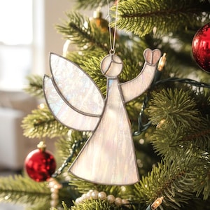 Pode incluir: Enfeite de Natal de anjo em vitral com asas iridescentes e um coração. O anjo está pendurado em uma árvore de Natal com enfeites vermelhos e luzes. O anjo é feito de vidro transparente com contorno branco.