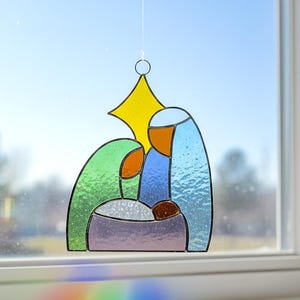 May include: Stained glass nativity scene with a yellow star, green, blue, orange, and purple hues. The artwork depicts Mary, Joseph, and baby Jesus. The piece is hanging in front of a window, with a clear blue sky in the background.