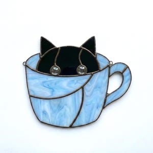 May include: A stained glass cat peeking out of a light blue teacup. The cat is black with two clear, round embellishments for eyes. The teacup has a handle and is made of blue and white swirled glass.