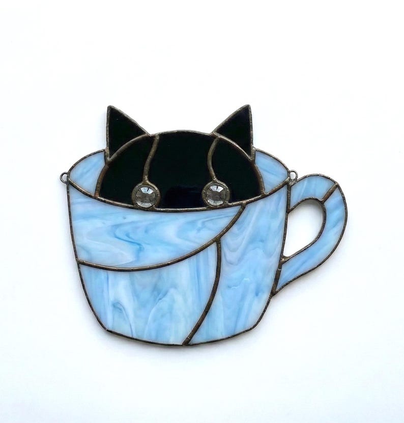 May include: A stained glass cat peeking out of a light blue teacup. The cat is black with two clear, round embellishments for eyes. The teacup has a handle and is made of blue and white swirled glass.