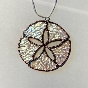 May include: A handcrafted pendant necklace with a sand dollar design. The pendant is made of iridescent glass with a copper-coloured border and a central five-petal flower design. The necklace has a silver-coloured chain.
