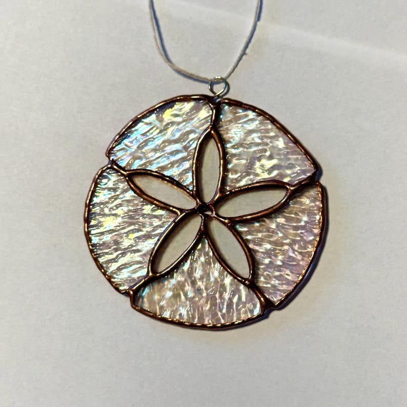 May include: A handcrafted pendant necklace with a sand dollar design. The pendant is made of iridescent glass with a copper-coloured border and a central five-petal flower design. The necklace has a silver-coloured chain.