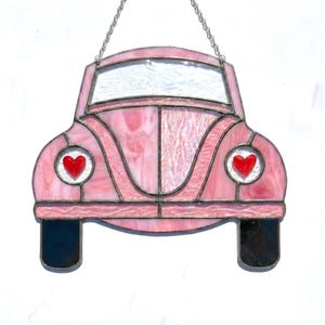 Op de afbeelding: Roze glas-in-lood auto met rode hartaccenten in de koplampen. De auto heeft zwarte banden en een zilveren ketting om op te hangen. Het glas heeft een gemarmerd effect. Een decoratief stuk voor Valentijnsdag of een autoliefhebber.