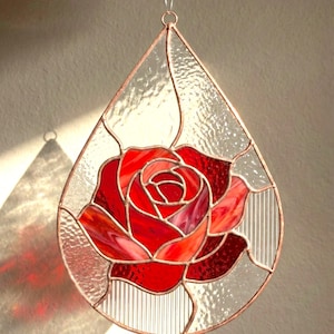 Può includere: Suncatcher a forma di goccia in vetro colorato con una rosa rossa. La rosa è composta da varie tonalità di vetro rosso, con vetro trasparente e strutturato intorno. Incorniciato in rame e con un anello per appenderlo.