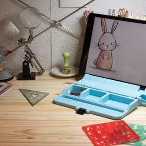 May include: A light blue pencil case holds an eraser. A tablet displays a bunny drawing. A ruler, triangle, and stencils are on a wooden surface. Other items include a magnifying glass, pencils, and a stamp.