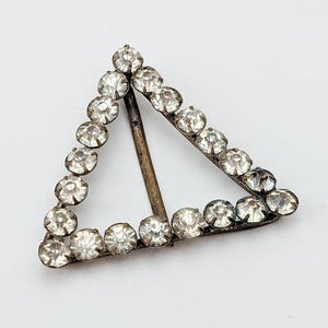 May include: A vintage triangular buckle, likely for a belt or accessory. The buckle is silver-toned and outlined with small, clear rhinestones. A central bar reinforces the triangle's shape. The buckle is set against a white background.