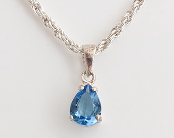 Könnte beinhalten: Eine silberfarbene Halskette mit einem tropfenförmigen, hellblauen Edelsteinanhänger. Die Kette hat ein gedrehtes Seildesign. Der Anhänger hat eine leuchtend blaue Farbe und ist in eine silberfarbene Metalleinfassung gefasst. Die Halskette wird vor weißem Hintergrund präsentiert.