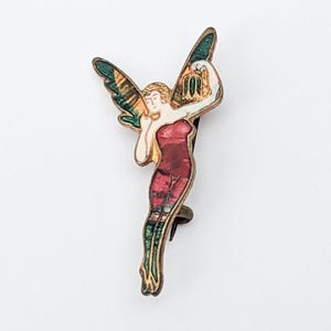 May include: A gold-toned pin with a colorful image of a fairy with green wings and a red dress.