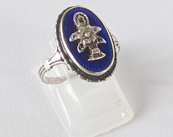 Könnte beinhalten: Silberring mit ovaler, dunkelblauer Emaille-Oberfläche und dekorativem Silberdesign. Der Ring hat einen silbernen Rand und eine kleine, detaillierte Silberfigur in der Mitte. Der Ring wird auf einem transparenten Ständer vor weißem Hintergrund präsentiert.