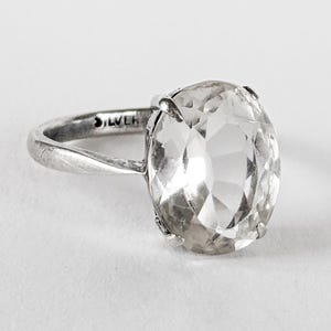 Op de afbeelding: Een zilveren ring met een ovale, heldere edelsteen. De smalle band is gegraveerd met "SILVER". De gefacetteerde edelsteen vangt het licht.
