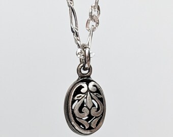 Könnte beinhalten: Eine silberne Halskette mit einem kleinen, ovalen Anhänger. Der Anhänger hat ein detailliertes, durchbrochenes Design mit floralem Muster. Die Kette ist eine filigrane Gliederkette. Der Hintergrund ist einfarbig hell.