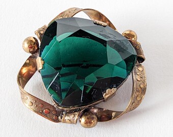 Könnte beinhalten: Vintage Brosche mit einem großen, facettierten smaragdgrünen Edelstein. Der Edelstein ist in einem goldfarbenen Metallrahmen mit dekorativen Details und kleinen, runden Akzenten gefasst. Die Brosche hat ein klassisches, elegantes Design.