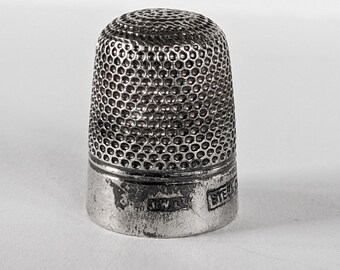 Könnte beinhalten: Ein silberner Fingerhut mit einer strukturierten Oberfläche. Der Fingerhut hat eine zylindrische Form mit einem leicht ausgestellten Boden. Das Metall hat ein stumpfes, gealtertes Aussehen. Der Fingerhut ist zum Nähen und Basteln bestimmt. Die Höhe beträgt ca. 2,5 cm.