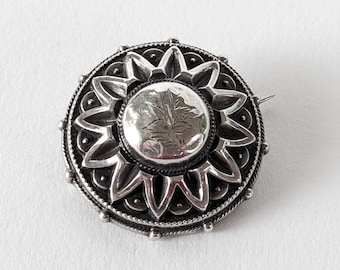 Könnte beinhalten: Detaillierte, silberfarbene Brosche mit kreisförmigem Design. Die Brosche hat ein zentrales, graviertes Medaillon, umgeben von sternförmigen Elementen und einem Perlenrand. Die Nadel ist vor einem weißen Hintergrund platziert.