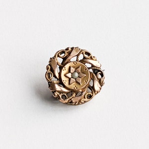May include: A detailed, antique gold-toned brooch. The brooch features a central star design with a small pearl-like bead, surrounded by an ornate, filigree pattern. The brooch is circular and set against a white background.