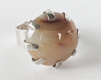 Könnte beinhalten: Ein Silberring mit einem großen, oval geformten, braunen und beigefarbenen Stein. Der Stein wird von silbernen Zinken gehalten. Der Ring ist ebenfalls aus Silber und hat ein Markenzeichen. Der Ring ist vor einem weißen Hintergrund platziert.