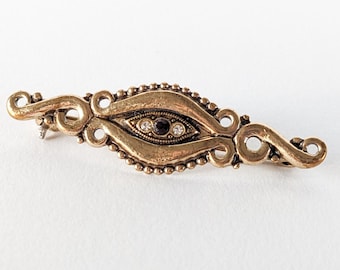 Könnte beinhalten: Eine verzierte Brosche im Vintage-Stil in Goldtönen. Die Brosche hat ein augenförmiges zentrales Design mit einem dunklen Edelstein und klaren Kristallen. Die Ränder sind mit kleinen Perlen und Wirbelmustern verziert, was eine antike Ästhetik erzeugt.