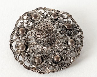 Könnte beinhalten: Silberfarbene Filigranbrosche mit floralem Design. Die Brosche hat ein zentrales, strukturiertes Element, umgeben von einem Muster aus Blumen und kleinen, runden Akzenten. Die filigrane Metallarbeit erzeugt eine zarte, Vintage-Ästhetik.