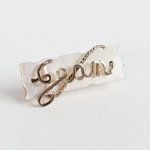 May include: A silver-toned wire name pin that reads "Sean" in cursive script. The name is attached to a rectangular, off-white, mother-of-pearl base with a textured edge. The pin is set against a white background.