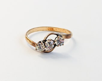 Könnte beinhalten: Ein goldener Ring mit drei klaren Steinen, die in einem geschwungenen Design gefasst sind.