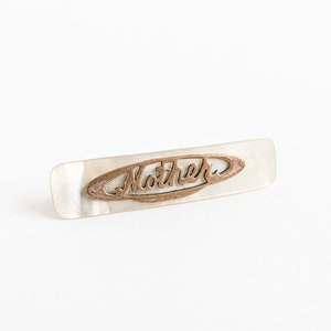 May include: A white mother of pearl barrette with a gold-toned metal inscription that reads "Mother".