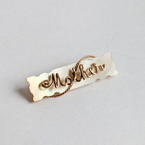 May include: A white mother of pearl brooch with a gold wire inscription that reads "Mother".
