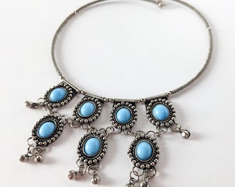 Könnte beinhalten: Silberfarbene Choker-Halskette mit ovalen, hellblauen Edelsteinen. Die Halskette hat eine filigrane Kette und kleine, baumelnde Glockenanhänger. Die Edelsteine sind in einem perlenbesetzten Rahmen gefasst, was einen Vintage-Look erzeugt.