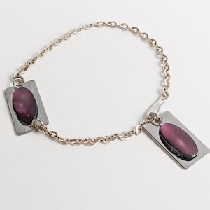 以下が含まれることがあります： シルバーカラーのブレスレット。チェーンと長方形のペンダントが特徴です。各ペンダントはシルバーフレームと、中央に楕円形の濃い紫色の石があります。ブレスレットは白い背景に表示されています。