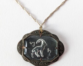 Könnte beinhalten: Eine Vintage-Halskette mit einem Anhänger, der das silberne Zodiakzeichen Steinbock zeigt. Der Anhänger ist in einem dekorativen, dunklen Metallrahmen gefasst und hängt an einer filigranen goldfarbenen Kette. Der Hintergrund ist weiß.