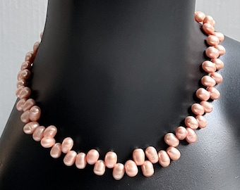 Könnte beinhalten: Eine pfirsichfarbene Perlenkette, die auf einer schwarzen Mannequin-Büste präsentiert wird. Die Halskette besteht aus kleinen, unregelmäßig geformten Perlen, die ein zartes und elegantes Design ergeben. Die Büste steht vor einem neutralen Hintergrund.