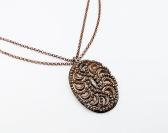 Könnte beinhalten: Eine Halskette aus Kupferkette mit einem großen ovalen Anhänger. Der Anhänger ist aus Metall mit einem filigranen Design und einer braunen Glasmitte.