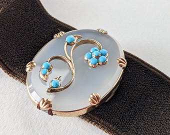 Könnte beinhalten: Eine ovale antike Brosche mit goldenem Rahmen und hellgrauer Mitte. Die Brosche hat ein goldenes Blumendesign mit türkisfarbenen Akzenten. Die Brosche ist auf einem dunkelbraunen Samtband angebracht.