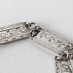 May include: Close-up of a silver bracelet with rectangular links. Each link features intricate engraved floral and geometric patterns. The bracelet is connected by small circular rings, creating a flexible design.
