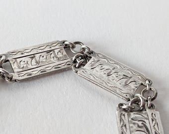 Könnte beinhalten: Nahaufnahme eines silberfarbenen Armbands mit rechteckigen Gliedern. Jedes Glied weist aufwendig gravierte Blumen- und geometrische Muster auf. Das Armband ist durch kleine kreisförmige Ringe verbunden.