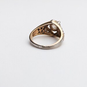 Op de afbeelding: Een zilveren ring met een grote, heldere edelsteen gezet in een gouden zetting. De ring is versierd met kleinere edelstenen rondom de zetting.