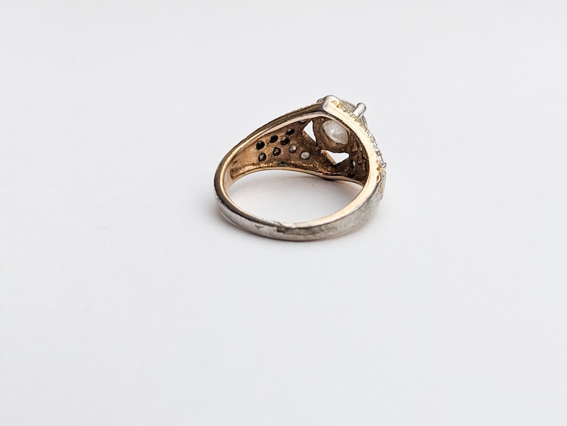 Op de afbeelding: Een zilveren ring met een grote, heldere edelsteen gezet in een gouden zetting. De ring is versierd met kleinere edelstenen rondom de zetting.