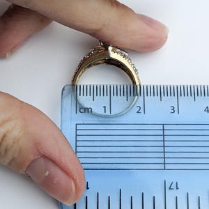 Op de afbeelding: Een gouden ring met een grote, heldere steen omringd door kleinere heldere stenen. De ring wordt gemeten met een liniaal en heeft een diameter van ongeveer 1,75 cm.