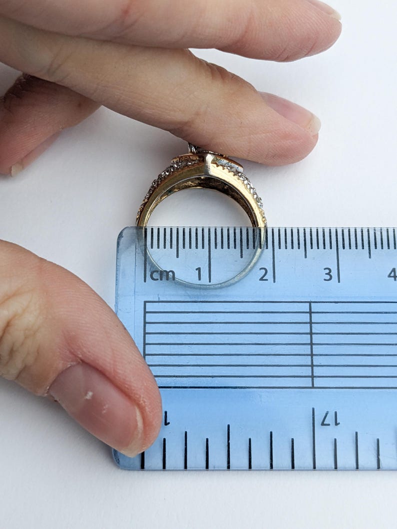 Op de afbeelding: Een gouden ring met een grote, heldere steen omringd door kleinere heldere stenen. De ring wordt gemeten met een liniaal en heeft een diameter van ongeveer 1,75 cm.