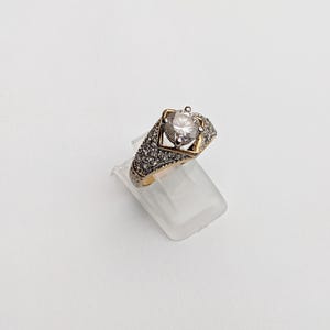 Op de afbeelding: Een gouden ring met een grote, heldere steen in een vierkant frame. De ring is versierd met kleinere, heldere stenen.