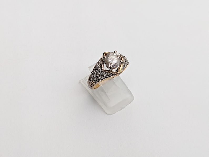 Op de afbeelding: Een gouden ring met een grote, heldere steen in een vierkant frame. De ring is versierd met kleinere, heldere stenen.