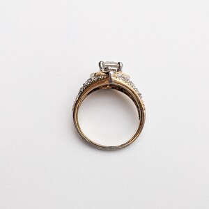 Op de afbeelding: Een gouden en zilveren ring met een grote, heldere steen in het midden. De ring is bezet met kleinere, heldere stenen rond de band.
