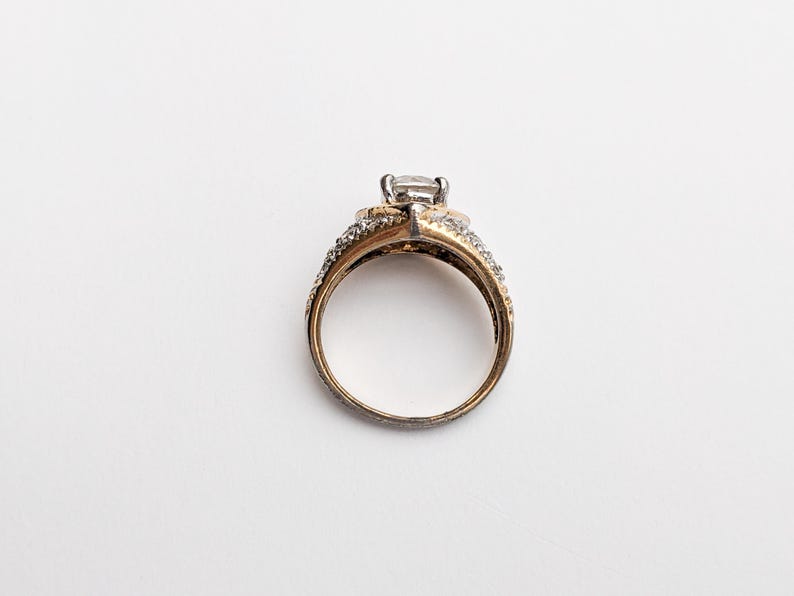 Op de afbeelding: Een gouden en zilveren ring met een grote, heldere steen in het midden. De ring is bezet met kleinere, heldere stenen rond de band.