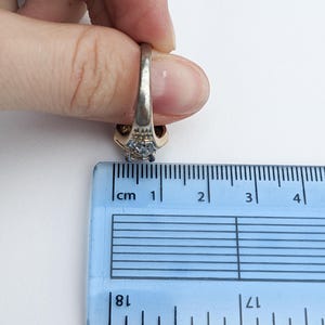 Op de afbeelding: Een gouden en zilveren ring met een blauwe edelsteen en kleinere heldere edelstenen. De ring wordt gemeten met een liniaal, wat aangeeft dat hij ongeveer 1,8 cm breed is.