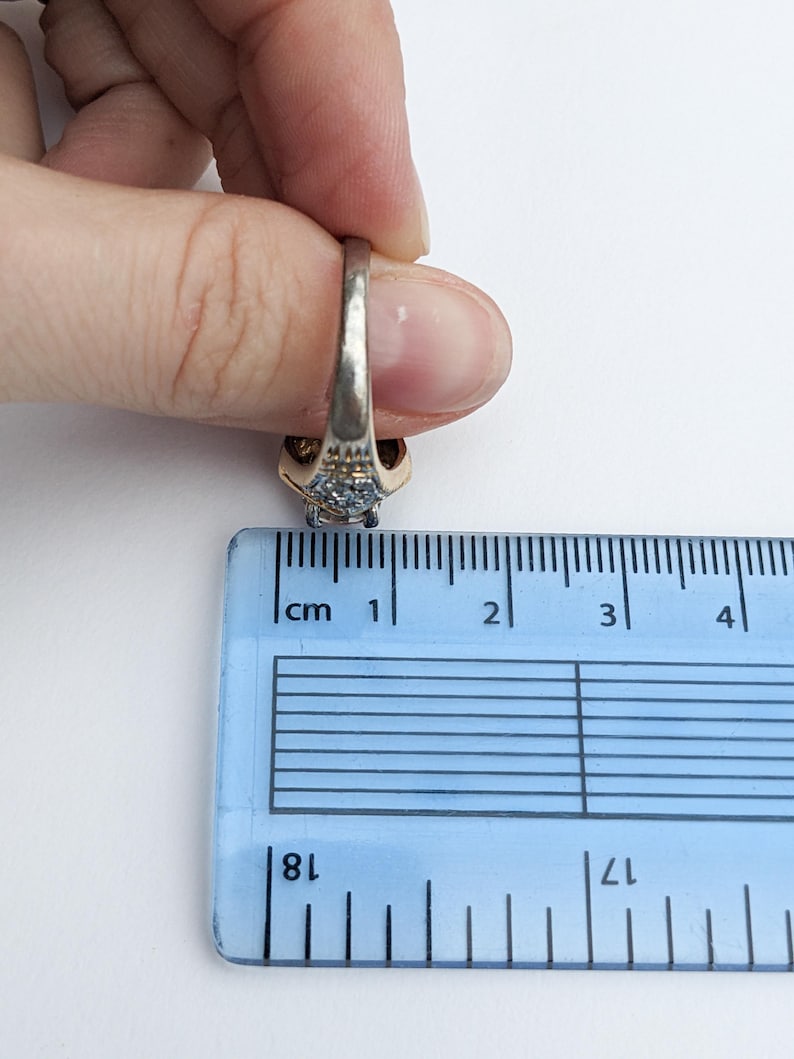 Op de afbeelding: Een gouden en zilveren ring met een blauwe edelsteen en kleinere heldere edelstenen. De ring wordt gemeten met een liniaal, wat aangeeft dat hij ongeveer 1,8 cm breed is.
